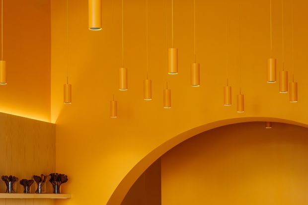 A dramatic monochromatic orange interior featuring a high ceiling with numerous slim pendant lights, a sweeping curved arch or vaulted opening, and warm integrated lighting. A small display shelf with decorative objects is visible at the lower left. The space has a bold, sculptural and highly coordinated fit-out.