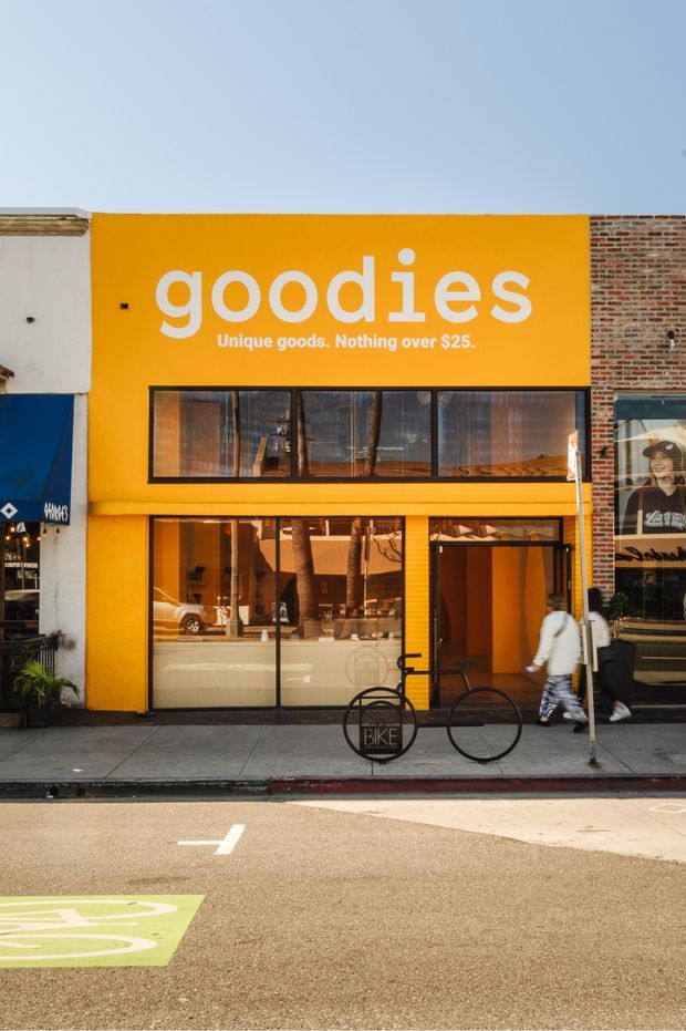 Street-facing storefront with a vivid orange painted facade, large white “goodies” signage, broad dark-framed glazing, and a brick-clad neighboring building. The shop fronts onto a paved urban sidewalk and marked street, with bicycles and pedestrians outside.