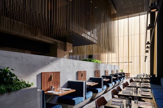 A sophisticated contemporary restaurant dining interior with a long, carefully set communal-style table in the foreground and several blue upholstered booth-style dining settings beyond. The space features a tall glazed facade with sheer drapery, a dramatic suspended ceiling installation of numerous slim timber or metal rods, textured grey concrete-like walls, integrated planters, dark ceiling panels, and warm ambient lighting. The palette combines charcoal, grey, timber brown, deep navy blue, and greenery, creating a refined, atmospheric hospitality setting.