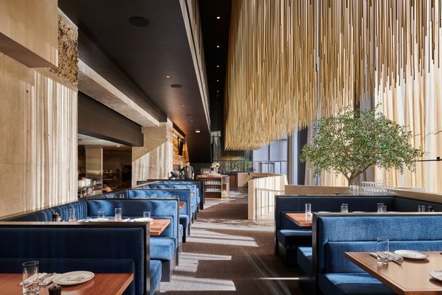 A sophisticated contemporary restaurant interior with rows of deep blue upholstered banquettes and armchairs arranged around timber-topped dining tables. The space features warm vertical timber wall panelling, a dramatic suspended installation of thin timber slats across the ceiling, dark ceiling surfaces with recessed spotlights, stone-textured structural elements, large windows, and a prominent indoor tree. The long, linear dining room has a refined, warm and atmospheric hospitality design.