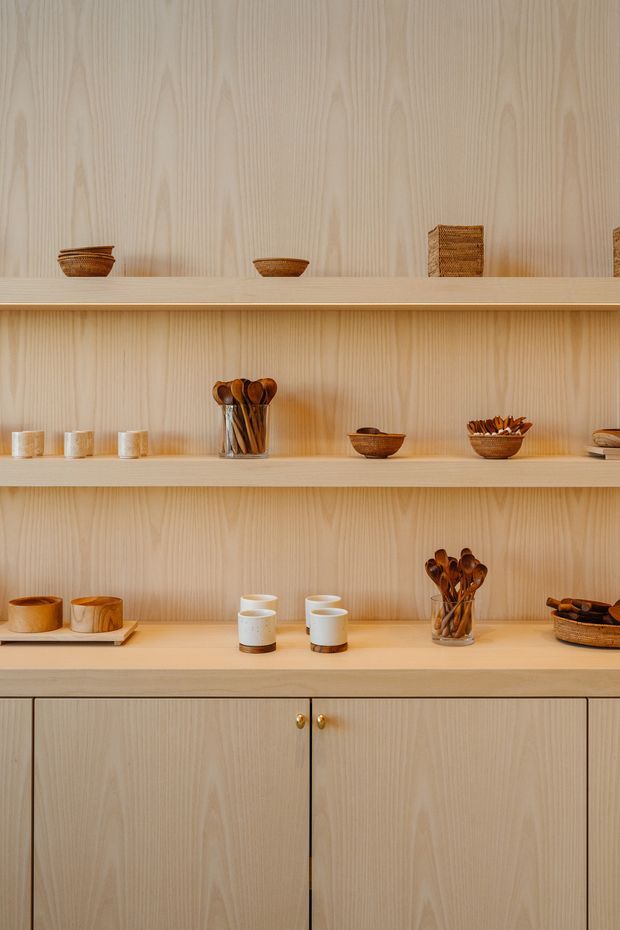A close-up interior view of a built-in light timber shelving and cabinetry unit. Three open display shelves hold ceramic cups, bowls, woven baskets, wooden utensils, and serving pieces, while the lower section has closed timber cabinet doors with small brass knobs. The design is warm, orderly, and minimalist, with strong vertical wood grain throughout.
