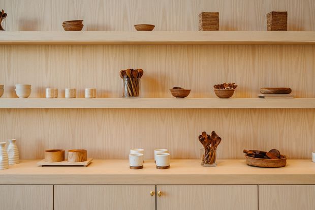 A warm, minimalist built-in display and storage wall with three long open timber shelves, pale vertical-grain wood panelling, and matching lower cabinetry. The shelves are styled with ceramic bowls, cups, small vessels, and wooden utensils, creating a refined natural presentation.