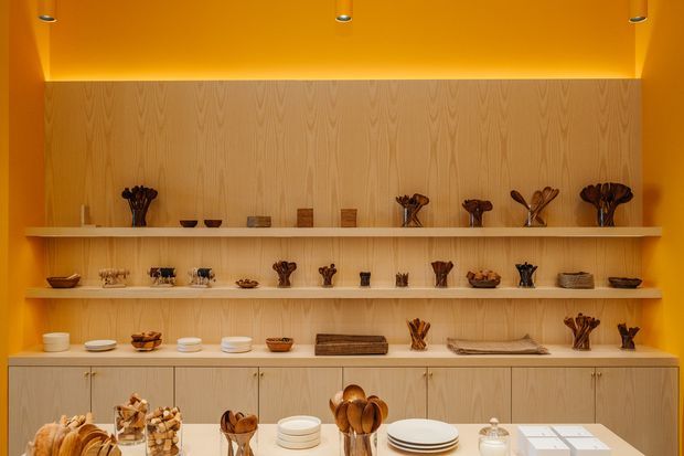 Warm, monochromatic display and storage wall with pale timber cabinetry, open shelves, and integrated lighting. The shelves present small decorative objects, ceramics, bowls, vessels, and tableware, while a central countertop displays additional pieces. Bright orange walls and ceiling create a bold, gallery-like interior.