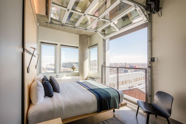 Bright compact bedroom with a platform bed, white bedding, dark accent cushions, and a striped throw. The room features exposed concrete columns, simple pale walls, expansive windows, and a distinctive overhead glazed opening with industrial-style framing and a raised mesh guard. A black molded chair sits near the window, with a broad urban cityscape visible outside.