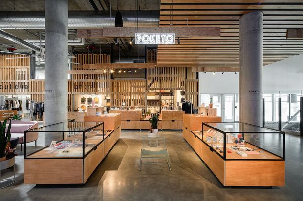 A contemporary commercial Pokéto retail and dining interior with a polished concrete floor, warm timber display counters, glass-topped merchandise cases, timber slatted ceiling and screening, exposed concrete columns, visible ductwork, and pendant lighting. The open-plan space combines a branded retail display area with a service counter and seating.