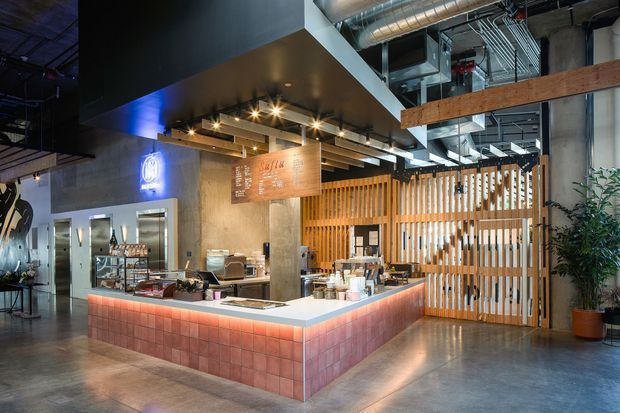 A contemporary café or hospitality counter in an industrial-style interior. The central service island has a tiled base with warm under-counter lighting, a pale stone or solid-surface benchtop, coffee equipment and display shelving. Behind it are timber vertical slats, exposed concrete columns, visible ductwork and services, dark ceiling elements, and a timber overhead beam. The space has a polished concrete floor, layered spot lighting, and a warm, refined industrial atmosphere.