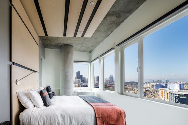 Bright contemporary bedroom in a high-rise apartment, featuring a bed positioned beside expansive corner windows with city views. The room has a minimalist built-in headboard wall, exposed concrete ceiling and cylindrical concrete column, black ceiling battens, and white bedding with a muted terracotta throw.