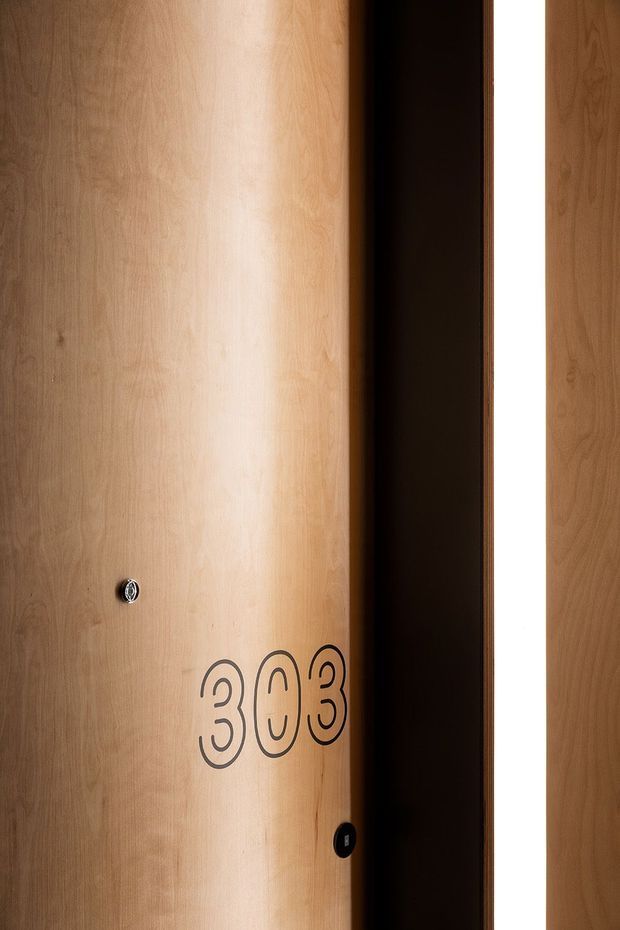 Close-up of a tall, curved timber veneer door or wall panel with a warm natural wood grain. The panel features dark outlined numerals reading “303” and small circular hardware or access fittings. A deep black vertical reveal separates it from a bright white opening, creating a high-contrast, minimalist architectural detail.
