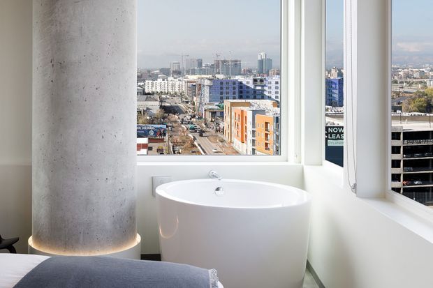 A bright contemporary bathroom with a freestanding white oval bathtub positioned beside large corner windows. A round raw concrete structural column is prominent in the foreground, with a cityscape visible outside. The lower edge of a bed is visible at the bottom left, suggesting the bathroom opens directly from or is integrated with a bedroom.
