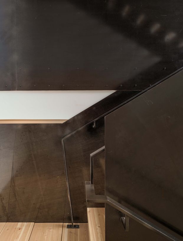 A close architectural view of a compact stairway with dark metal wall and balustrade surfaces, timber stair treads, and a contrasting white geometric opening or ceiling plane. The composition emphasizes angular lines, exposed construction, and a dark industrial material palette.