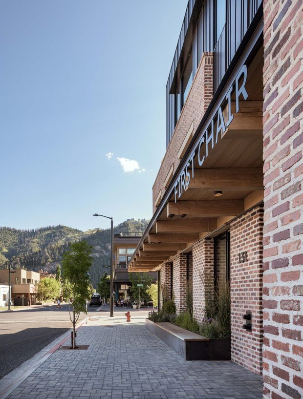 Street-level architectural view of a contemporary commercial building in a mountain town. The facade combines exposed red brick, dark metal-framed glazing, and a deep timber-lined canopy with large projecting signage. A paved pedestrian walkway, landscaped planter beds, street trees, and neighbouring low-rise buildings lead toward forested mountains in the background.