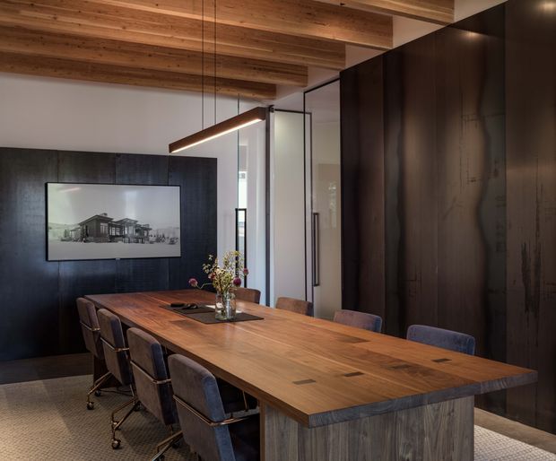 A refined dining room or meeting space centered on a long solid timber table with upholstered chairs. The room features dark wood or black built-in wall panelling and cabinetry, exposed timber ceiling beams, a suspended linear pendant light, and glazed partitions or doors at the rear. The palette is warm timber, charcoal, and muted grey, creating a sophisticated contemporary atmosphere.