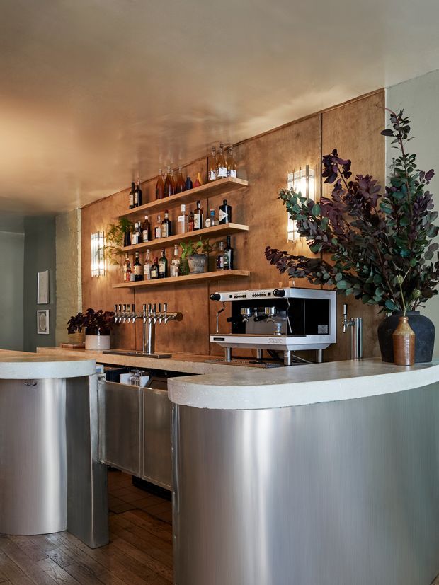 A compact, warmly lit bar area with curved stainless-steel counters, a white solid-surface countertop, exposed wooden flooring, open timber liquor shelves, beer taps, and a commercial espresso machine. The backdrop is a textured warm brown wall with decorative wall sconces, creating an intimate hospitality-style setting.