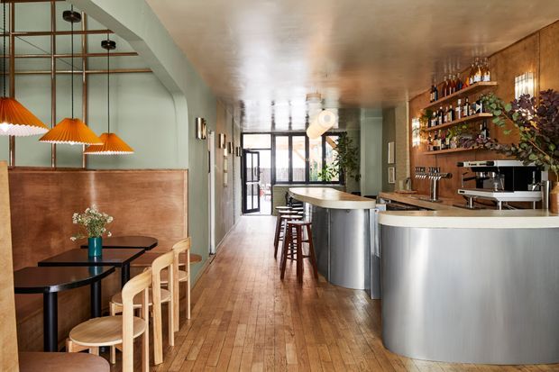 A stylish narrow hospitality bar and dining space with warm timber flooring, curved pale-green walls, copper-toned ceiling, timber booth panelling, black tables, and light timber stools. A long curved bar with brushed metal cladding, timber shelving, bottles, glassware, and coffee equipment occupies the right side, while pendant lights and a glazed entry brighten the room.