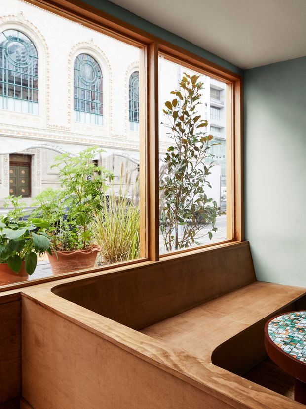 A bright interior nook with large timber-framed windows overlooking a historic ornate building and planted courtyard. A built-in timber bench wraps around the corner beneath the windows, paired with a small round mosaic-topped table. The room has pale green walls, warm timber joinery, and abundant natural light.