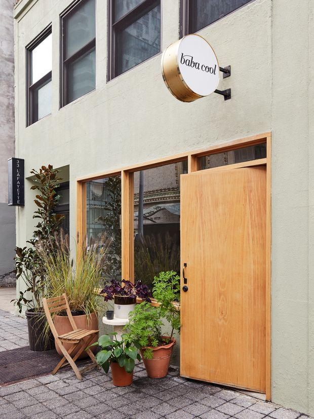 Street-level view of a small commercial storefront with a pale green rendered facade, dark-framed upper windows, warm timber-framed glazing, and a large natural timber entrance door. A round projecting sign reads “baba cool.” Potted plants and a folding timber chair soften the paved frontage.