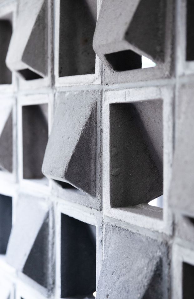 Close-up architectural detail of a grid-like concrete breeze-block screen or perforated masonry wall. The rough light-grey concrete blocks have deep rectangular openings and angled, projecting faces that create strong geometric shadows and texture. The image is monochromatic and abstract, focused on the fixed wall construction rather than a room or broader setting.