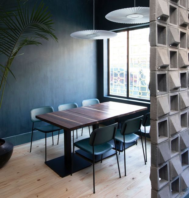 A compact dining area with a dark blue textured wall, pale timber flooring, and a rectangular dark timber dining table surrounded by modern teal upholstered chairs. Sculptural white pendant lights hang above the table, while a large black-framed window provides natural light. A distinctive exposed concrete breeze-block partition forms the right side of the image, adding a strong mid-century and industrial architectural feature.
