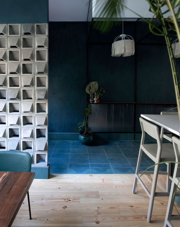 A moody, eclectic dining area with deep blue walls, blue tiled flooring, pale timber floorboards, a white dining table and chairs, and a distinctive breeze-block partition. A fabric pendant light, potted plants, and dark framed wall panels add layered texture and contrast.