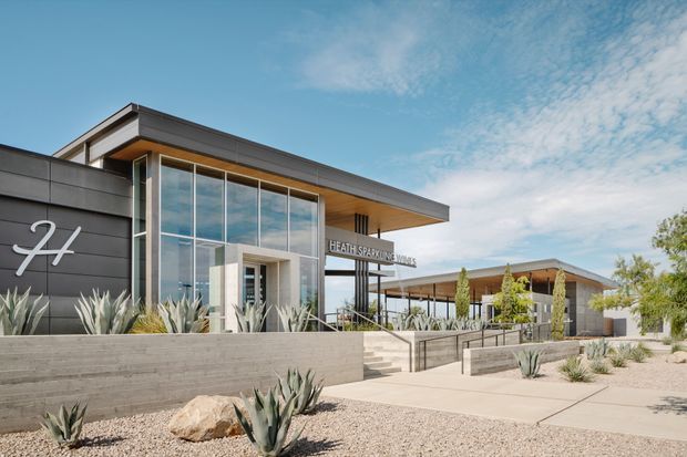 A contemporary low-rise commercial or public building with a bold dark panelled facade, extensive floor-to-ceiling glazing, projecting flat roof planes, and a covered entry walkway. The entrance signage reads “Heath Sparkling Wines,” indicating a winery or wine-related commercial venue. Drought-tolerant agave and other architectural planting sit within a gravel landscape, with concrete retaining walls, steps, and accessible-looking pathways.