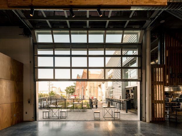 A contemporary industrial-style commercial interior opening onto an outdoor terrace through a large raised glazed garage-style door. The space features exposed roof framing and services, warm timber wall cladding, a polished concrete floor, dark metal-framed glazing, and café-style tables and chairs. An urban laneway and adjacent brick and corrugated-metal buildings are visible beyond the terrace.