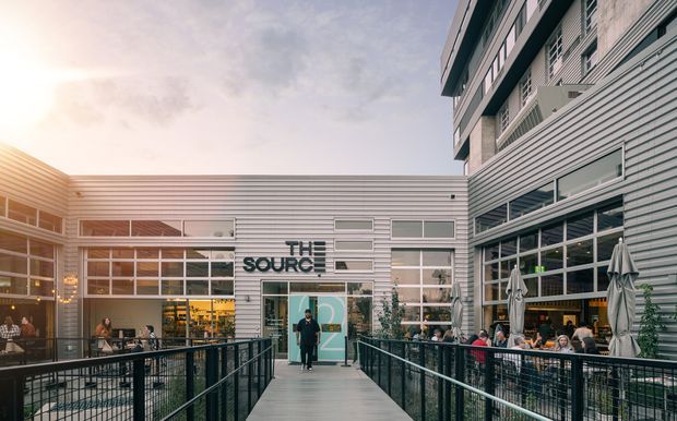 Exterior view of a contemporary multi-storey commercial or mixed-use building arranged around an open pedestrian courtyard. The facade features horizontal metal cladding, extensive grid-pattern glazing, exposed concrete or masonry upper levels, black metal railings, outdoor dining tables, and umbrellas. A central walkway leads toward a glazed entrance with visible signage reading “THE SOURCE.”