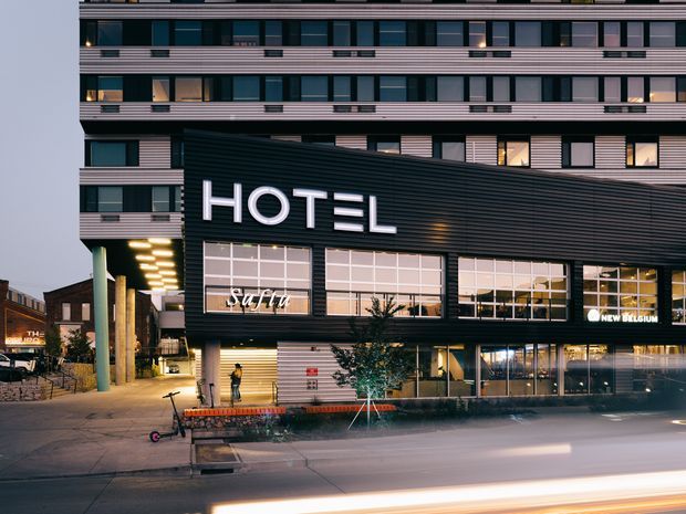Exterior dusk view of a multi-storey contemporary hotel building in an urban streetscape. The facade combines dark horizontal metal cladding, extensive rectangular glazing, illuminated signage reading “HOTEL” and “Sofitel,” and lighter banded upper-storey cladding. Street-level entrances and hospitality spaces are visible, with blurred traffic light trails in the foreground.