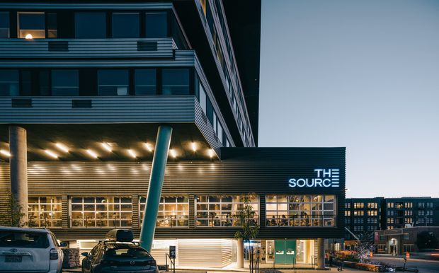 Dusk exterior view of a contemporary multi-storey commercial building with dark metal cladding, extensive glazing, projecting horizontal volumes, and dramatic angled structural columns. The ground level contains a brightly illuminated retail or hospitality tenancy with visible signage reading “The Source,” while surrounding urban buildings and parked vehicles frame the scene.