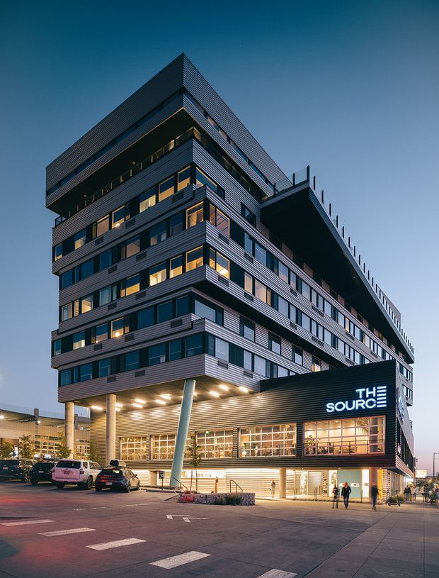 Dusk exterior photograph of a contemporary multi-storey mixed-use commercial building. The angular structure has extensive horizontal metal cladding, projecting balconies and terraces, large glazed windows, a covered ground-level entry, and illuminated signage reading “THE SOURCE.” The building fronts a paved urban forecourt with parked vehicles and pedestrians.