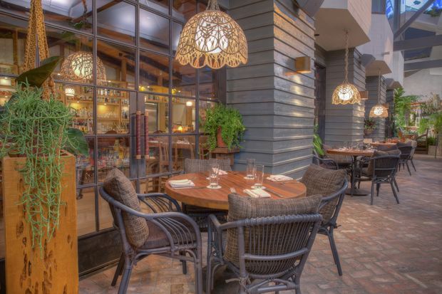 A warmly lit outdoor dining terrace or courtyard restaurant with round wooden tables, woven chairs, place settings, hanging woven pendant lights, abundant tropical plants, exposed brick paving, and a glass-fronted bar or dining area beside dark horizontal wall cladding. The space has a relaxed, atmospheric hospitality setting with strong indoor-outdoor connection.