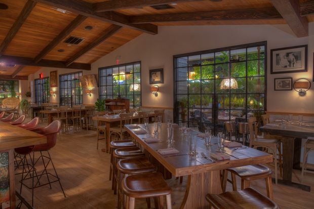 A warmly lit upscale restaurant dining room with numerous timber tables and leather seating, including a long communal table in the foreground. Large black-framed multi-pane windows overlook lush greenery, while exposed timber ceiling beams, wood panelling, framed artwork, wall sconces, and pendant lighting create a refined rustic atmosphere.