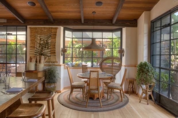 Warm, eclectic dining area with a round wooden dining table and woven cane chairs, positioned beneath a large suspended pendant light. The room features expansive black-framed windows and glazed doors, a timber-lined ceiling with exposed beams, pale timber flooring, white plaster walls with timber dado trim, indoor plants, woven wall art, and layered natural-fibre rugs. A kitchen or living area is partially visible at the left edge.