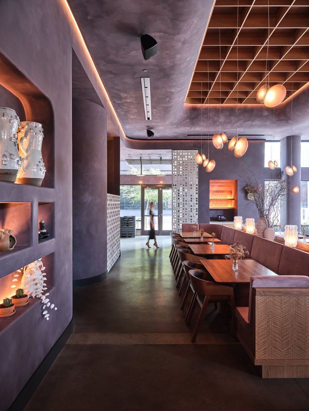 A sophisticated contemporary restaurant or hospitality dining space with a long banquette and rows of dark timber dining tables and chairs. The interior features deep mauve and charcoal plaster walls, dark polished flooring, warm integrated LED strip lighting, a dramatic timber slatted ceiling grid, sculptural pendant lights, built-in display niches, and a perforated decorative screen near the entrance. The atmosphere is intimate, warm, and refined.