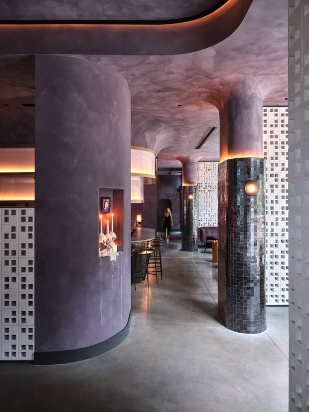A dramatic, moody commercial interior with a curved bar or lounge area, sculptural rounded columns and ceilings, dark purple textured plaster surfaces, black mosaic-tiled columns, exposed concrete flooring, warm concealed lighting, and perforated breeze-block screens. Bar stools and a counter are visible deeper in the space, with urban buildings seen through the screen openings.