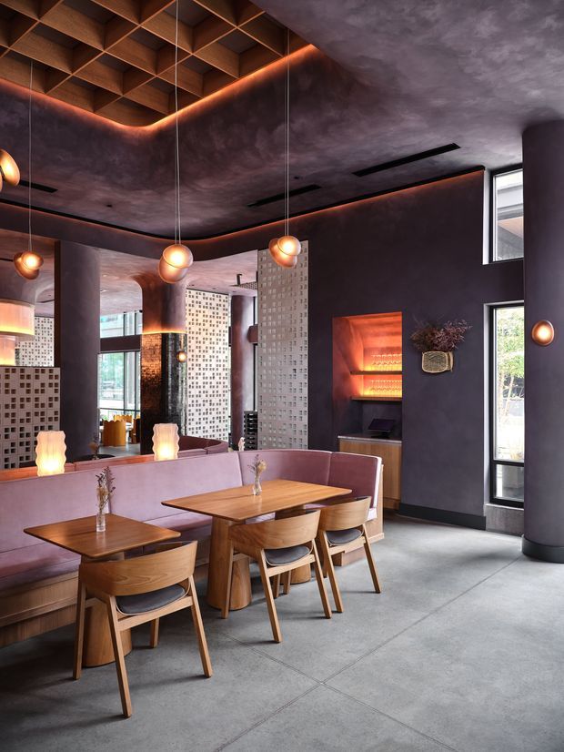 A moody, contemporary hospitality-style dining area with banquette seating, timber dining tables and chairs, dark plaster walls, glowing pendant lights, large mirrors, and decorative perforated masonry screens. A striking timber coffered ceiling with concealed amber lighting adds warmth, while tall windows bring in natural light. The space appears refined and atmospheric, with a built-in illuminated display niche and polished concrete flooring.
