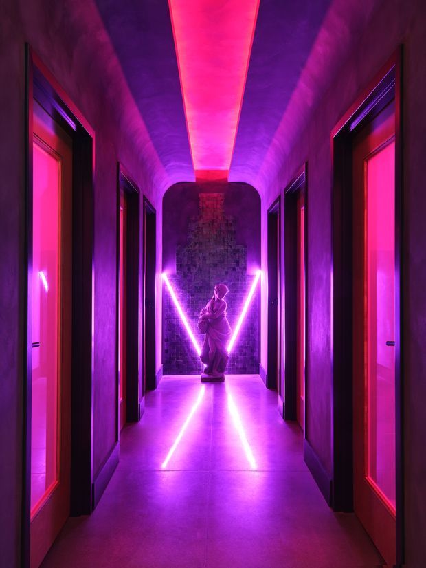 A dramatic, symmetrical interior corridor with multiple dark-framed doors on both sides, a curved ceiling, and a polished smooth floor. Saturated magenta and violet LED lighting creates a strong central axis, leading to a sculptural figure and textured dark feature wall at the far end. The atmosphere is theatrical, immersive, and highly moody.