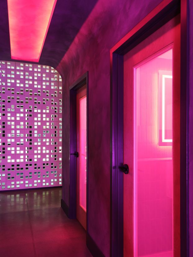 A dramatic, neon-lit interior corridor with dark purple walls and trim, vivid pink-red lighting, and several dark-framed doors with translucent or glowing pink panels. At the end of the corridor is a perforated decorative screen composed of many square openings, with a reflective dark floor and a curved ceiling soffit.