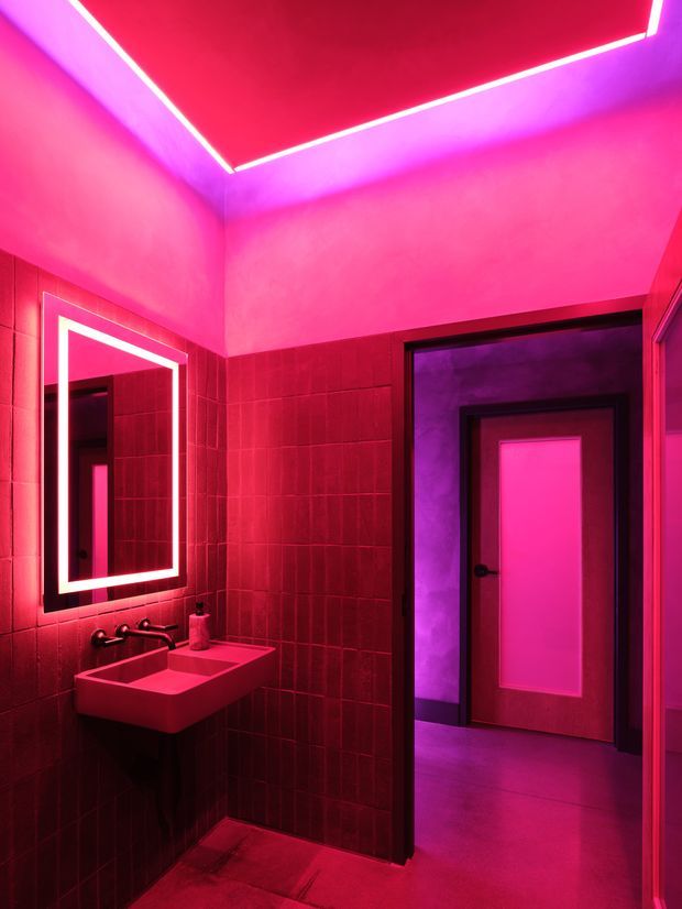 A compact, dramatically lit bathroom or powder room with dark red tiled walls and floor, a wall-mounted rectangular basin, black tapware, and a large illuminated mirror. Strong magenta and red LED lighting runs around the ceiling and mirror, while an open doorway reveals a connected corridor or adjacent space with a paneled door.