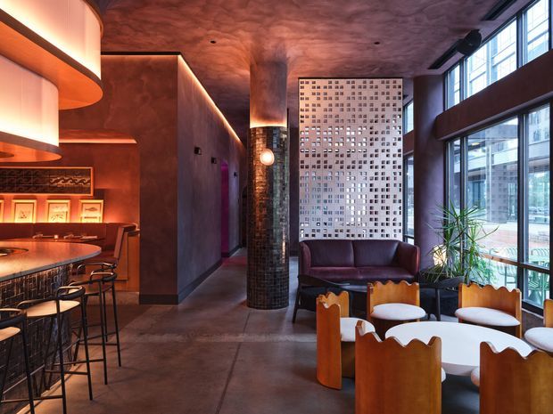 A dramatic, moody hospitality-style interior combining a bar and lounge seating area. The space features dark textured plaster or concrete-like walls and ceiling, polished dark flooring, warm integrated amber lighting, a curved bar with stools, leather lounge seating, sculptural timber chairs, large industrial-style steel-framed windows, and a perforated decorative screen. A tiled cylindrical column forms a strong central architectural feature.