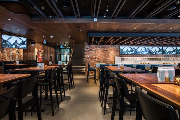 A warmly lit commercial dining and bar interior with exposed red-brick walls, extensive dark timber ceiling and wall lining, exposed dark ceiling beams, black-framed glazing, and rows of timber dining tables with black chairs and bar stools. A long bar counter with illuminated shelving and multiple wall-mounted screens runs along the rear wall, while a stairway leads to an upper level at the centre. The atmosphere is contemporary, moody, and industrial-inspired.