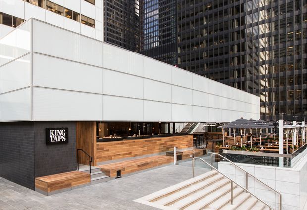 Exterior view of a contemporary commercial hospitality venue in a dense urban high-rise setting. A projecting white-panelled upper volume sits above a dark tiled facade and timber-clad stepped entry seating, with the King Taps sign visible. Outdoor dining terraces with umbrellas and tables occupy the adjacent paved plaza, accessed by broad stairs and ramps.