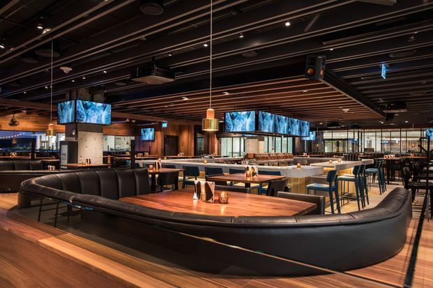 A spacious, dimly lit contemporary commercial dining and bar venue with extensive booth seating, dark leather banquettes, timber-topped tables, bar stools, pendant lights, and multiple wall-mounted screens. The ceiling features exposed dark services and linear timber slats, while large glazed openings brighten the rear of the space. The atmosphere is polished, warm, and moody.