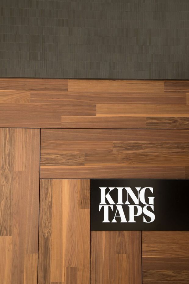 Close-up of a branded display wall featuring varied walnut-toned timber joinery and panel sections arranged in a geometric composition. A dark textured vertical panel spans the upper portion, while a black inset sign reading “KING TAPS” appears prominently among the wood panels. The image is primarily a material and retail branding detail rather than a complete room view.