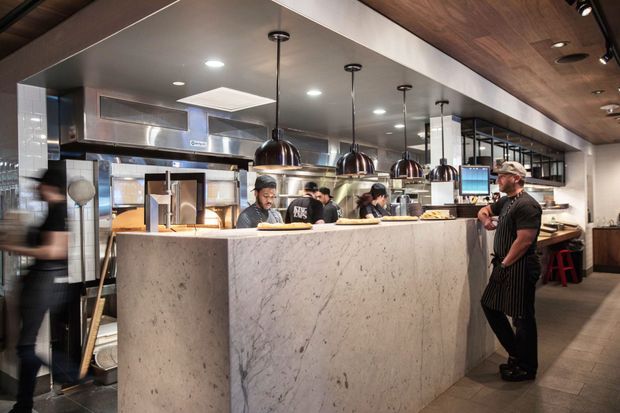 A contemporary commercial restaurant kitchen and service counter, viewed from the dining or customer area. A large pale stone-fronted counter separates customers from chefs working in an open stainless-steel kitchen. Black pendant lights hang beneath a dark timber ceiling, with stainless-steel equipment, shelving, bar stools and warm task lighting contributing to a polished hospitality fit-out.