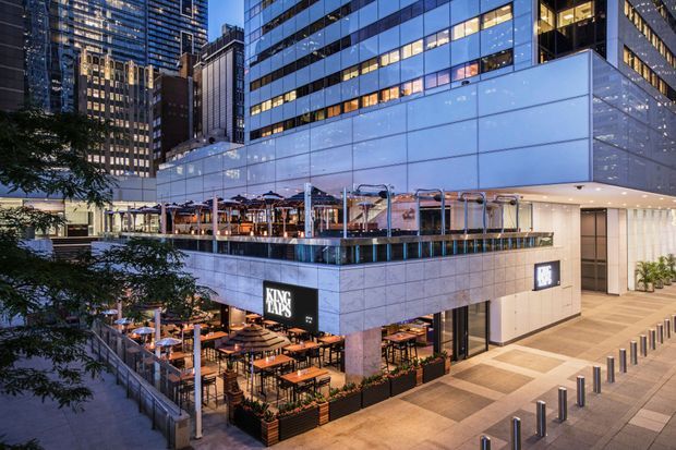 Evening exterior view of a multi-level urban commercial building, with a glazed upper volume, illuminated office towers in the background, and a restaurant with outdoor dining terraces at ground and upper levels. A paved pedestrian forecourt with bollards occupies the right side.