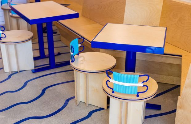 Playful contemporary interior featuring pale plywood wall panelling and built-in bench seating, square tables with vivid blue painted edges and legs, and cylindrical plywood stools. Curved blue floor inlay lines echo the bold blue accents. A bright turquoise accordion-like object sits on a foreground stool, adding a strong graphic accent.