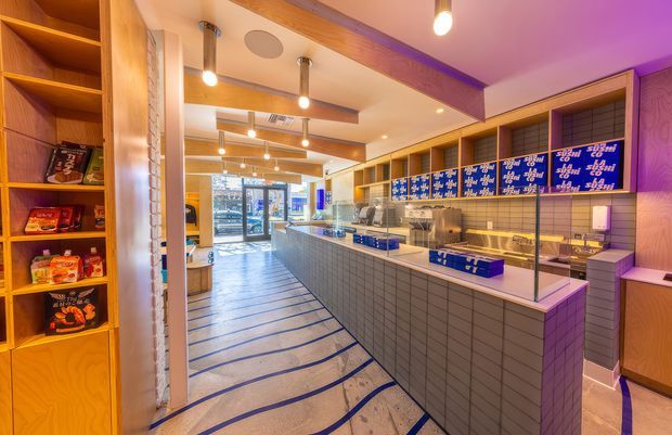 Interior view of a contemporary commercial food-service retail space, likely a bakery, takeaway, or dessert shop. The long tiled service counter has a glazed display and blue-branded storage boxes, with warm timber shelving and cabinetry, white tiled walls, exposed timber ceiling beams, pendant and recessed lighting, and a patterned floor with blue linear accents. Purple accent lighting adds a vivid contemporary atmosphere.