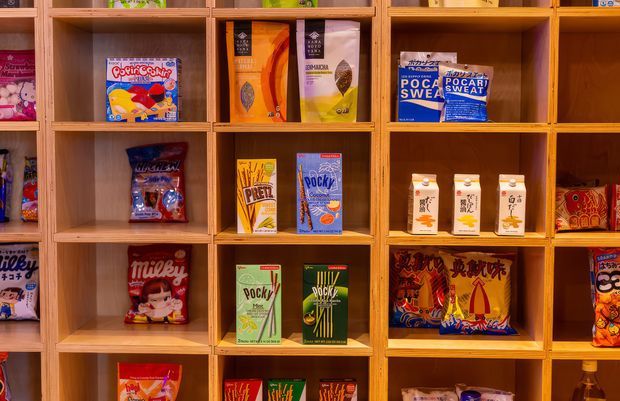 A close-up view of a warm timber display shelving unit arranged in a grid of square compartments, stocked with colourful packaged snack products, including Japanese confectionery and biscuits. The shelves dominate the frame and create a retail or commercial interior setting.