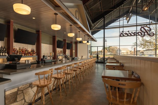 A spacious, warmly lit bar or restaurant interior with a long linear counter, timber stools, booth seating along the windows, and a large glazed facade. The space combines exposed industrial roof structure and services with a timber-lined ceiling, stone-faced bar base, dark metal framing, and pendant lighting. Bottles and bar equipment are arranged behind the counter, while a prominent reversed script sign appears on the glazing.