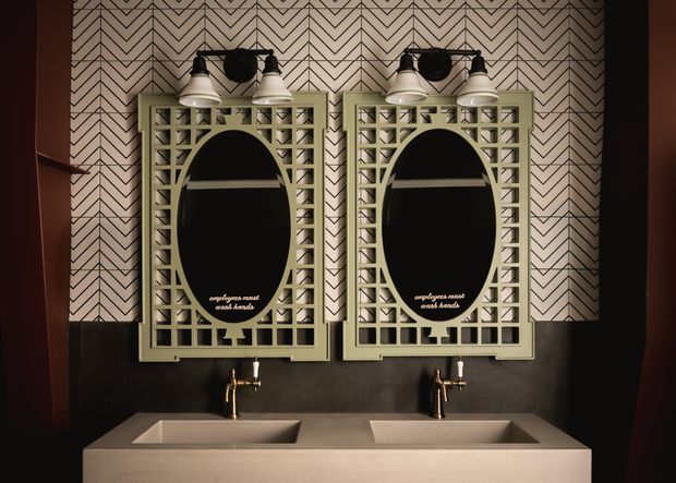 A dramatic, symmetrical bathroom vanity featuring two rectangular vessel sinks, brass wall-mounted taps, and twin oval mirrors set within pale green geometric openwork frames. The wall behind is covered in a bold black-and-off-white zigzag patterned finish, with dark lower wall panelling and paired industrial-style wall lights. The composition has a refined vintage/art-deco-inspired character with warm, moody lighting.